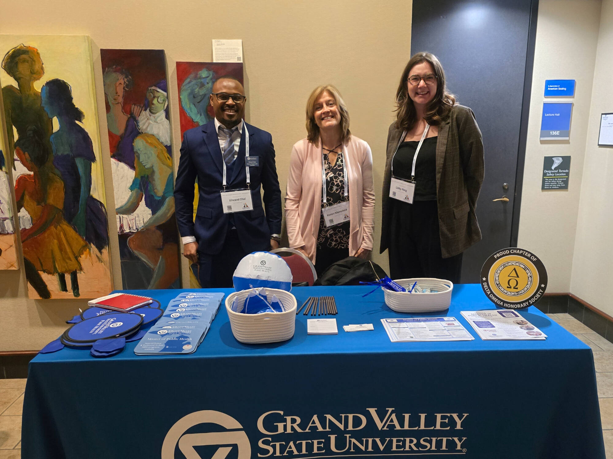 A current MPH student, a faculty member, and an MPH alumna pose, smiling behind a table displaying information about GVSU's MPH program.